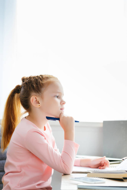 side view of thoughtful schoolgirl holding pen and looking away while studying at home - Photo, Image
