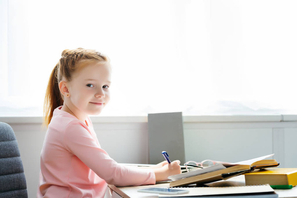side view of beautiful schoolgirl smiling at camera while writing in notebook at desk - Zdjęcie, obraz