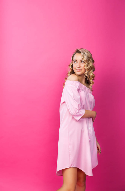 Pretty stylish girl with long curly hair wearing beautiful pink dress and posing against pink background. Fashion vogue style portrait of young happy smiling woman, copy space. - Photo, Image