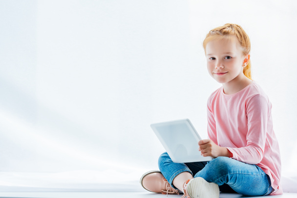 beautiful redhead child using digital tablet and smiling at camera while sitting on windowsill - Photo, image