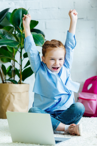 cheerful little schoolgirl raising hands while studying with laptop at home - Photo, Image