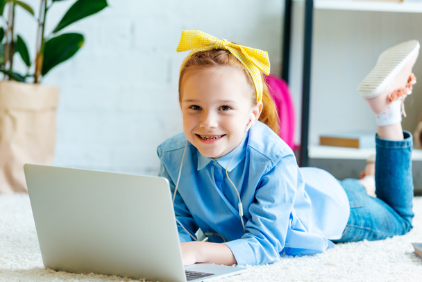 entzückendes kleines Kind mit Kopfhörern, das in die Kamera lächelt, während es auf dem Teppich liegt und Laptop benutzt  - Foto, Bild