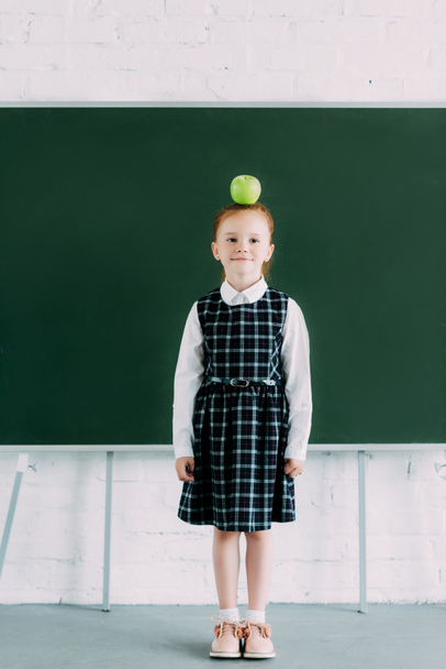 full length view of beautiful little redhead schoolgirl with apple on head looking at camera - Photo, Image