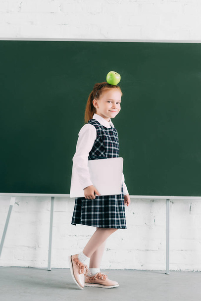 belle petite écolière avec pomme sur la tête tenant ordinateur portable et souriant à la caméra
  - Photo, image