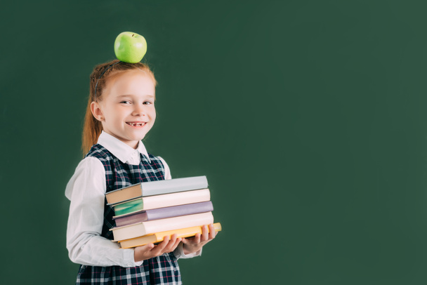 belle petite écolière avec pomme sur la tête tenant pile de livres et souriant à la caméra
  - Photo, image