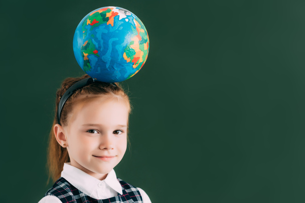 adorable little schoolchild with globe on head smiling at camera while standing near blackboard - Photo, Image