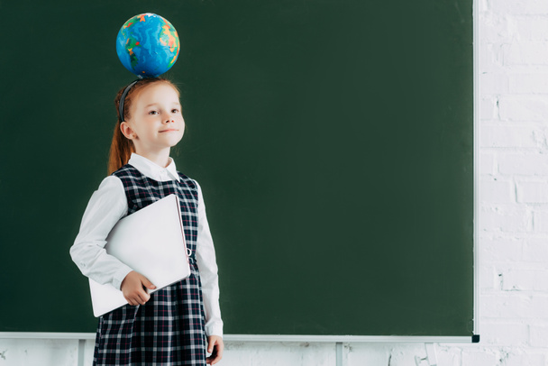 beautiful little schoolgirl with globe on head holding laptop and standing near chalkboard - Фото, изображение