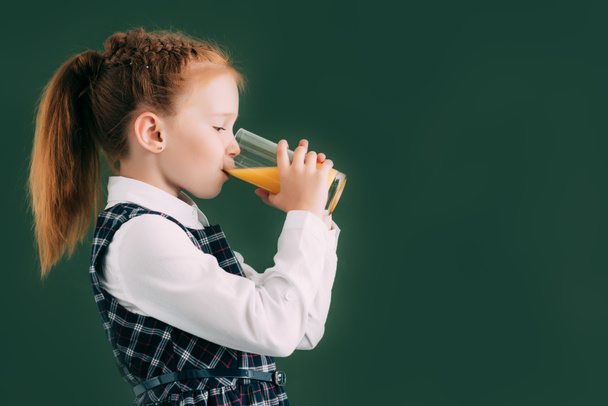 side view of adorable little schoolgirl drinking orange juice while standing near chalkboard - Fotó, kép