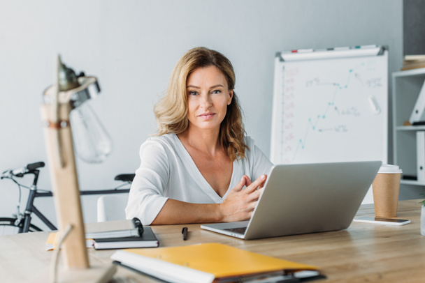 attraktive Geschäftsfrau sitzt mit Laptop am Tisch im Büro und blickt in die Kamera  - Foto, Bild