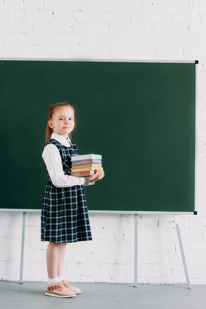 entzückendes kleines Schulmädchen, das einen Stapel Bücher in der Hand hält und in die Kamera schaut, während es neben der Tafel steht - Foto, Bild