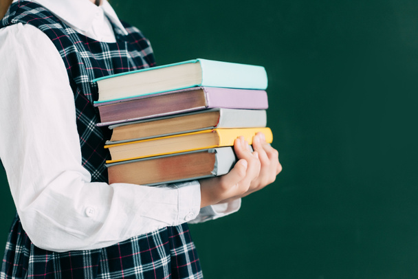 cropped shot of schoolgirl holding pile of books while standing near blackboard - Foto, Imagen