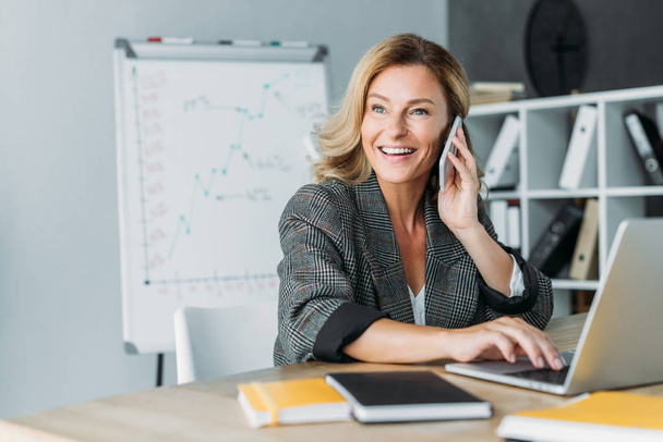 smiling beautiful businesswoman talking by smartphone in office and looking away - Photo, Image