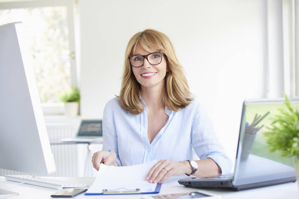 Знімок привабливої бізнес-леді середнього віку, що працює на комп'ютері та ноутбуці, сидячи за офісним столом
.  - Фото, зображення