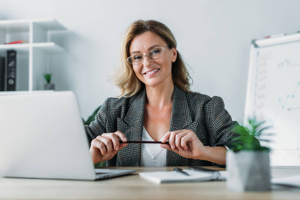 улыбающаяся привлекательная деловая женщина сидит за столом в офисе и смотрит в камеру
 - Фото, изображение