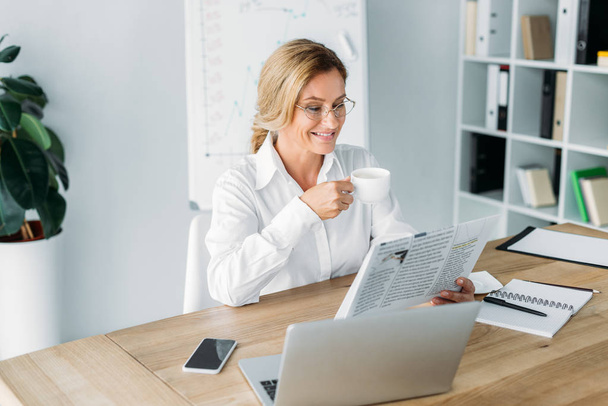 smiling attractive businesswoman drinking coffee and reading newspaper in office - Фото, изображение