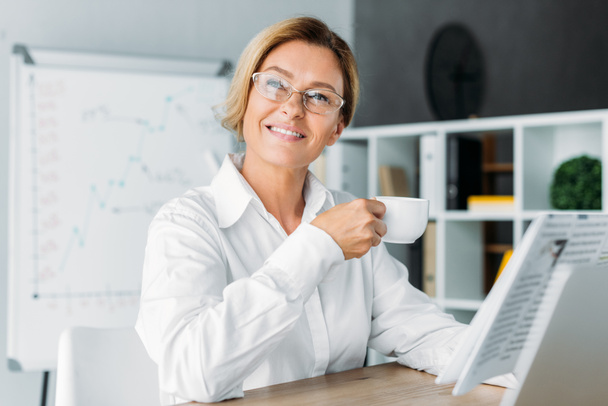 atractiva mujer de negocios sosteniendo la taza de café y mirando hacia otro lado en la oficina
 - Foto, imagen