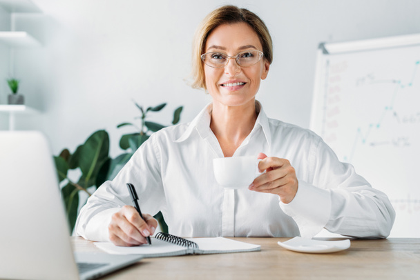 atractiva empresaria sosteniendo una taza de café y escribiendo a un cuaderno en la oficina
 - Foto, Imagen