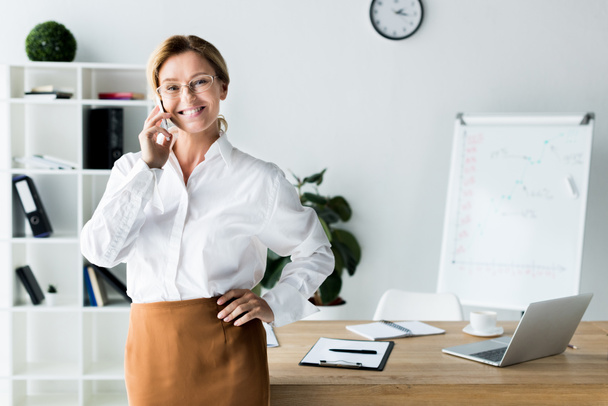 smiling attractive businesswoman talking by smartphone in office - Photo, Image