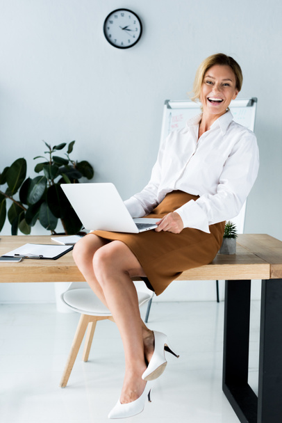 atrakcyjna kobieta siedzi na stole z laptopa i śmiejąc się w biurze - Zdjęcie, obraz