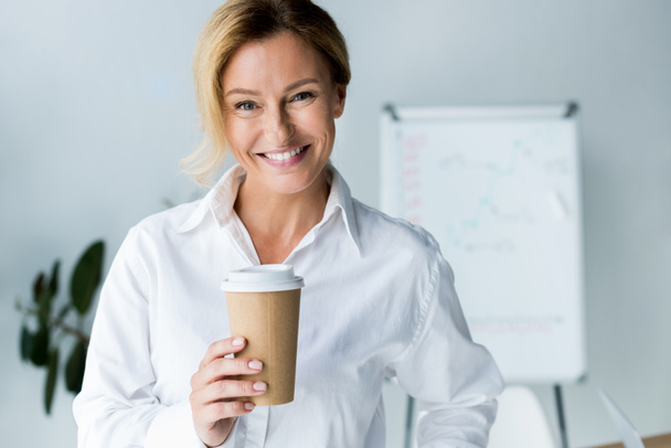 lachende aantrekkelijke zakenvrouw holding wegwerp koffiekopje in office en de camera kijken - Foto, afbeelding