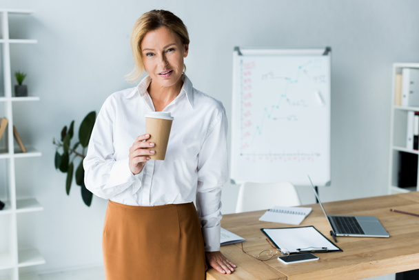 beautiful businesswoman holding disposable coffee cup in office and looking at camera - Fotografie, Obrázek