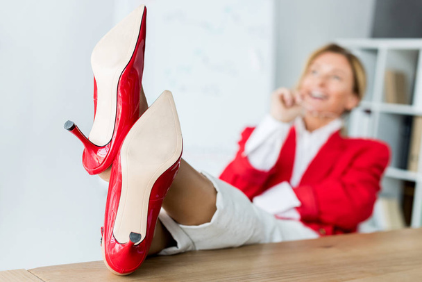 attraente donna d'affari ridente e seduta con le gambe sul tavolo in ufficio con le scarpe rosse in primo piano
 - Foto, immagini