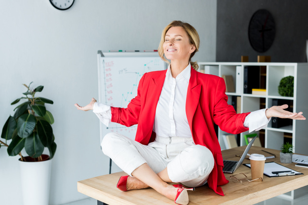 счастливая привлекательная деловая женщина медитирует в позе лотоса на столе в офисе
 - Фото, изображение