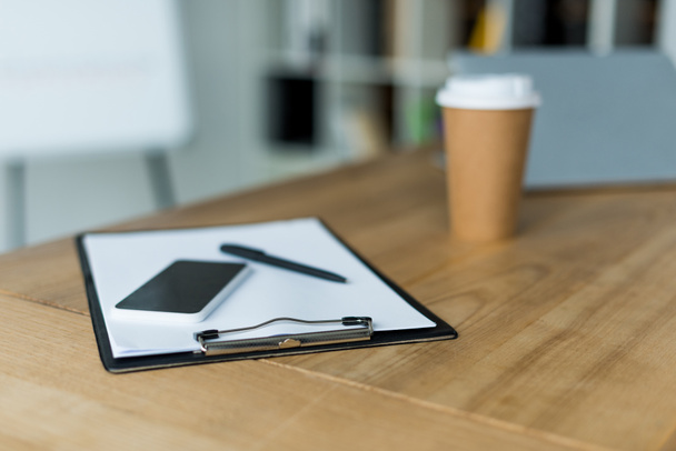 Smartphone und Einweg-Kaffeetasse mit Klemmbrett auf Holztisch im Büro - Foto, Bild