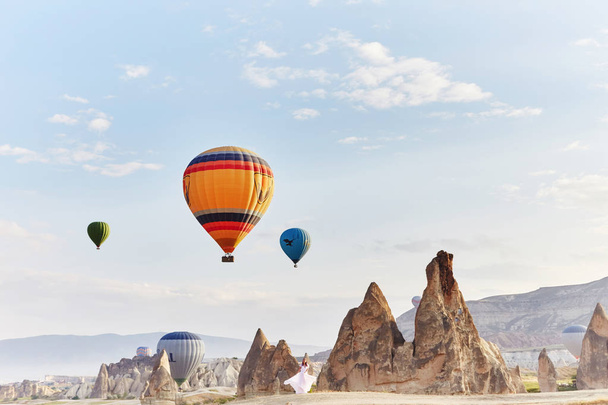 Woman in a long dress on background of balloons in Cappadocia. Girl with flowers hands stands on a hill and looks at a large number of flying balloons. Turkey Cappadocia fairytale scenery of mountains - Photo, image