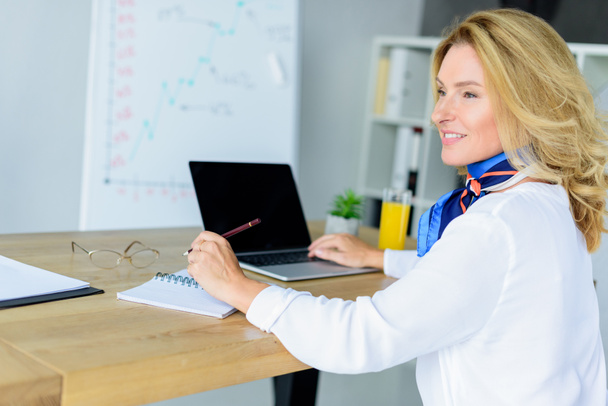 smiling attractive businesswoman sitting at table with laptop in office - Photo, Image
