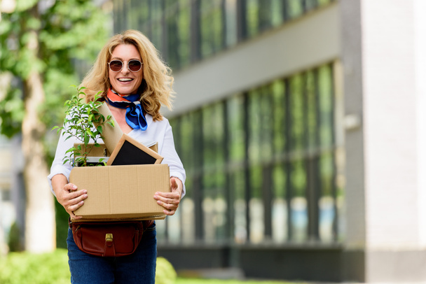 усміхнена приваблива жінка тримає паперову коробку з офісними речами на вулиці
  - Фото, зображення