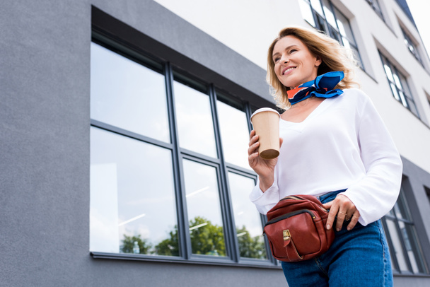 низький кут зору привабливої жінки, що йде з одноразовою чашкою кави на вулиці
  - Фото, зображення