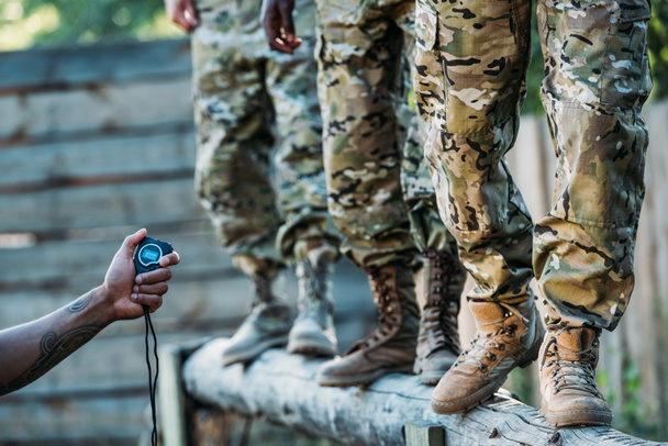 vista parcial del instructor táctico con stop watch examinando soldados multirraciales durante carrera de obstáculos en el rango
 - Foto, Imagen