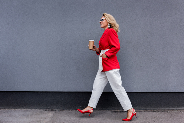 vista lateral de mujer atractiva en chaqueta roja caminando con café en taza de papel en la calle
  - Foto, imagen