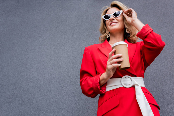 attractive woman in red jacket holding coffee in paper cup and touching sunglasses on street  - Фото, изображение
