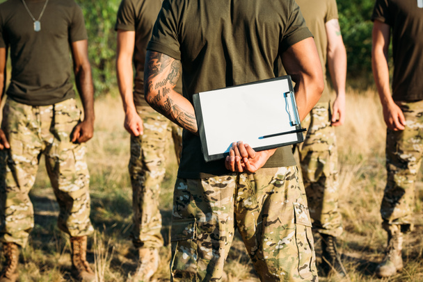 vista parcial del instructor táctico con bloc de notas y soldados multirraciales en uniforme militar en rango
 - Foto, Imagen