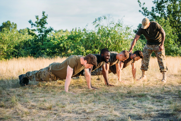 tactical instructor examining multicultural soldiers in military uniform doing push ups on range - Fotó, kép