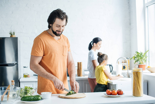 onnellinen nuori isä leikkaamalla kurkkua salaattia, kun hänen vaimonsa ja tyttärensä tiskaavat keittiössä
 - Valokuva, kuva