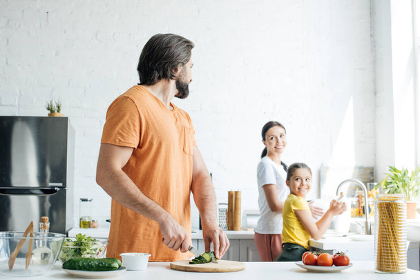ハンサムな若い父は彼の妻と娘が台所で皿洗いしながらサラダのため切削キュウリ - 写真・画像