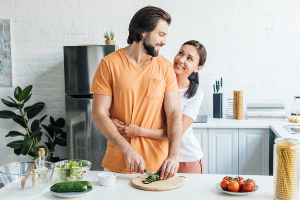 Kobieta, obejmując jego męża, podczas gdy on przygotowanie Sałatka razem w kuchni - Zdjęcie, obraz