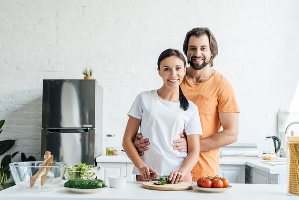 guapo sonriente hombre abrazando a su esposa mientras ella preparando ensalada juntos en la cocina
 - Foto, imagen
