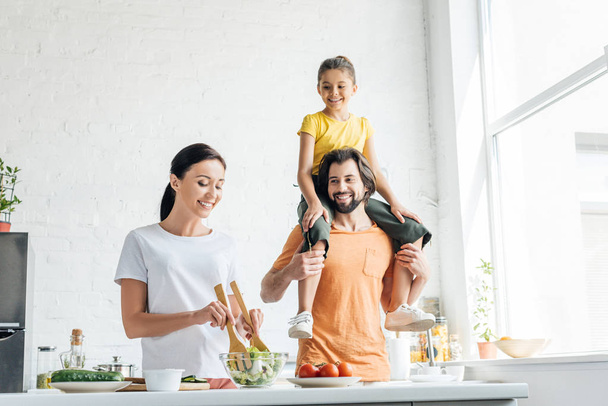 smiling young woman preparing salad while her daughter riding on shoulders of husband at kitchen - Foto, Imagen