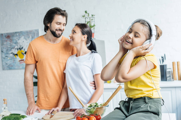 lachende meisje in hoofdtelefoons zittend op tafel terwijl haar ouders omarmen op achtergrond op keuken - Foto, afbeelding