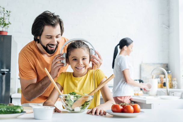 父と一緒に料理をお皿を洗う母がキッチンで背景にぼやけながらヘッドフォンで幸せな娘の笑顔 - 写真・画像
