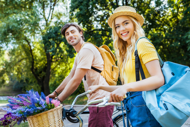 vista lateral de pareja sonriente con bicicletas mirando a la cámara en el parque de verano
 - Foto, Imagen