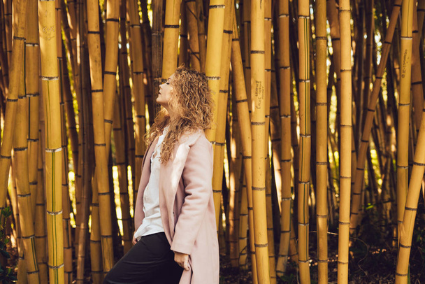curly stylish woman in coat posing next to tall bamboo trees in park - Foto, afbeelding