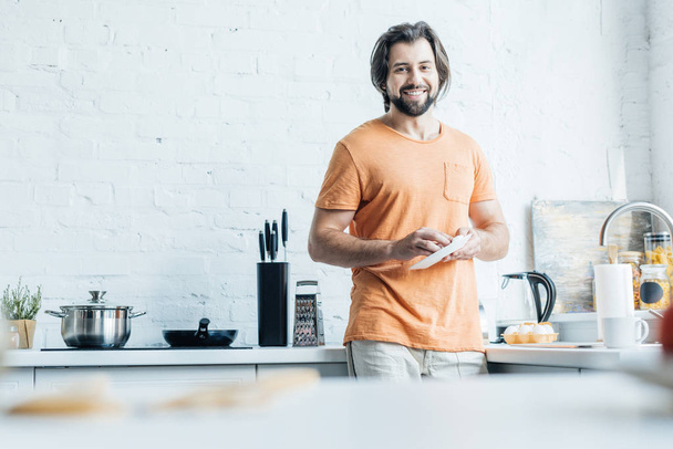handsome bearded man washing dishes alone at kitchen and looking at camera - Photo, Image