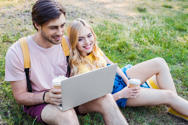 joven sonriente pareja con café para ir usando el ordenador portátil en el parque
 - Foto, Imagen