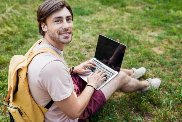 vista lateral do homem sorridente com mochila e laptop sentado na grama verde no parque
 - Foto, Imagem