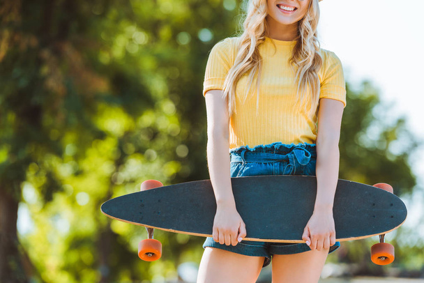 częściowy widok uśmiechający się blond kobieta z longboard stojąc na ulicy - Zdjęcie, obraz